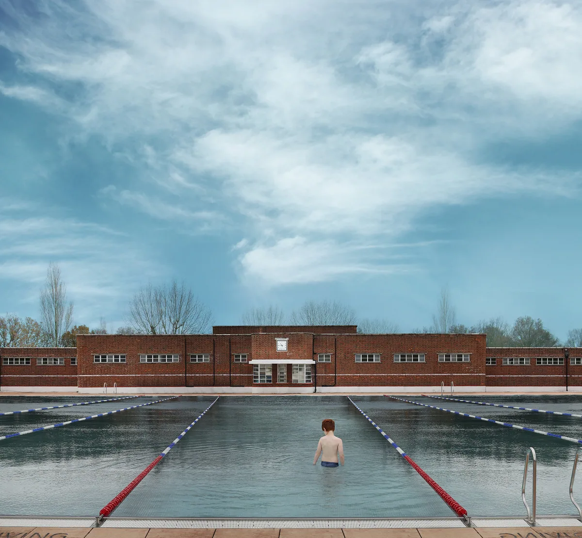 Photograph of a lone swimmer in Parliament Hill Lido beneath a wide, cloudy blue sky, the brick lido buildings beyond.