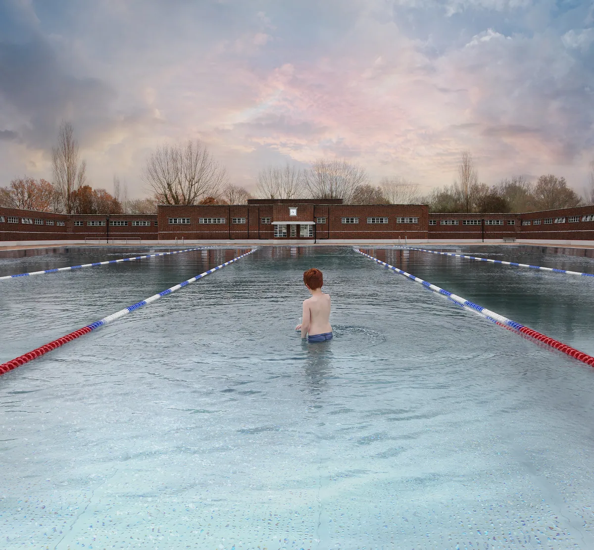 Photograph of a swimmer standing waist-deep in Parliament Hill Lido under a soft pink sky, lane ropes reaching towards the lido building.