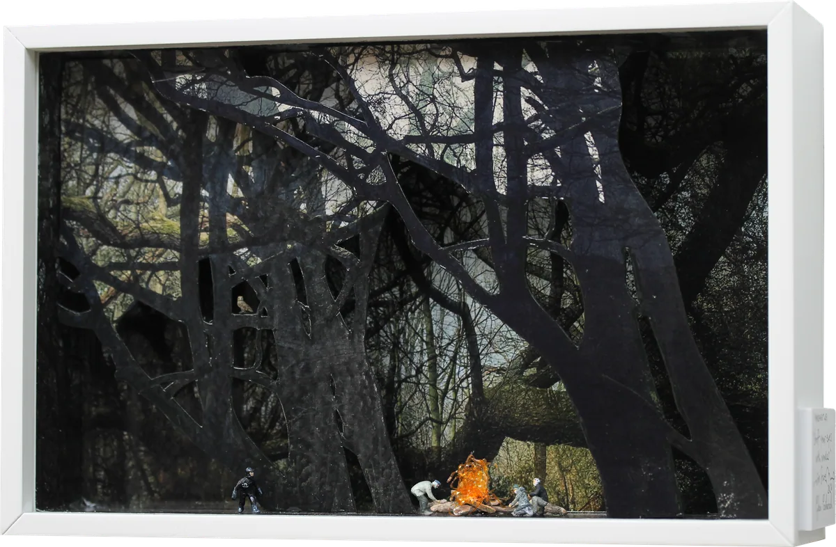 White box diorama of black silhouetted trees, with tiny figures gathered round a glowing campfire.