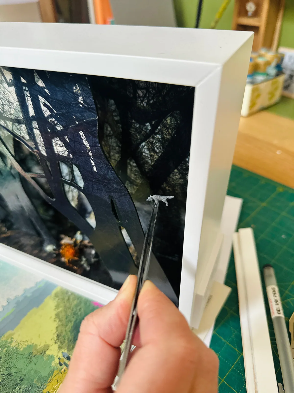 In the studio: tweezers placing a tiny white wolf among the dark cut-out trees, the campfire glowing below.