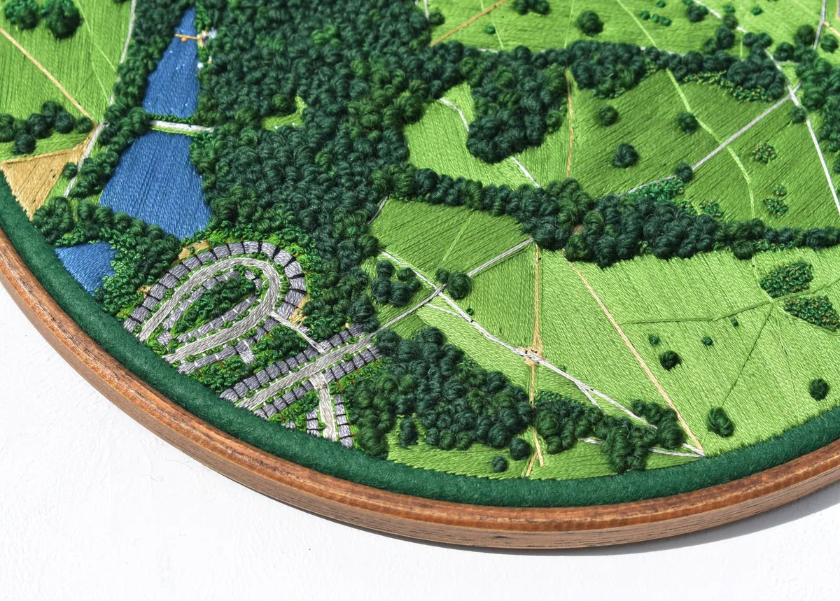 Detail: stitched rows of houses beside meadows and a pond, at the edge of the hoop.